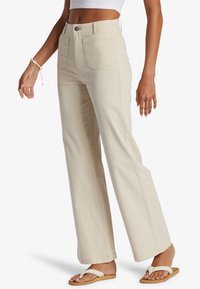 Frau in hoch taillierten, cremfarbenen, weit geschnittenen Hosen, weißem Crop-Top, geflochtenen Sandalen und einem Muschelarmband am linken Handgelenk, die seitlich steht.