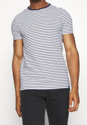 Homme portant un t-shirt blanc avec de fines rayures noires horizontales et un pantalon noir, debout devant un fond blanc uni.