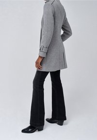 Cappotto grigio a spina di pesce con silhouette sagomata, maniche a imbuto e spacchi sul retro, abbinato a jeans neri a zampa d'elefante e stivali neri con tacco.