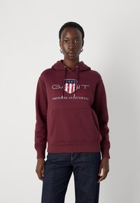 Felpa bordeaux con cordini, dotata di tasca frontale e di un logo con righe rosse e bianche. Il materiale sembra morbido e di peso medio.