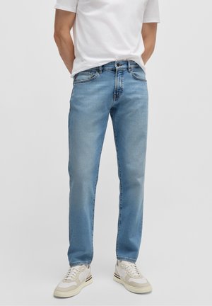 Mann trägt hellblaue Jeans mit geradem Schnitt, weißes T-Shirt und weiße Sneaker und steht vor einem unifarbenen Hintergrund.