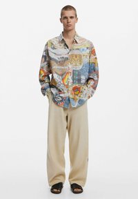 Colorful patterned button-up shirt featuring various graphics, paired with beige loose-fitting trousers and black slide sandals.