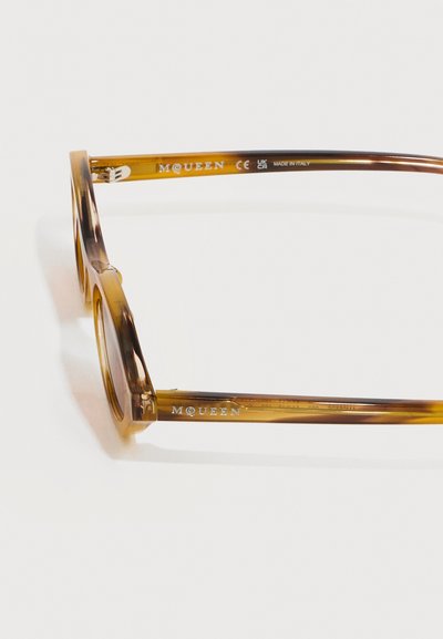 Καφέ διαφανή πλαίσια γυαλιών με στρογγυλούς φακούς, με την επωνυμία «McQueen» και την επιγραφή «Made in Italy» στα μπράτσα.