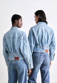 Dos personas llevan chaquetas de mezclilla azul claro con un diseño estampado, con costuras visibles y etiquetas de cuero de marca en la cintura.