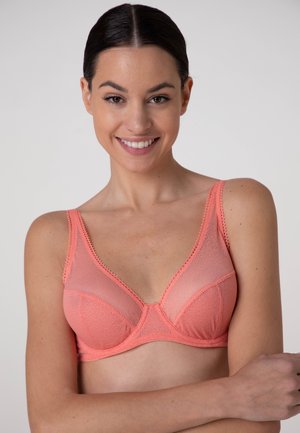Donna sorridente con capelli scuri che indossa un reggiseno rosa corallo trasparente, in piedi davanti a uno sfondo chiaro e uniforme con le braccia incrociate sotto il petto.