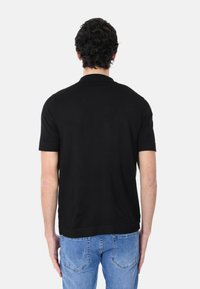 Polo shirt nero a maniche corte con colletto classico, caratterizzato da una texture liscia e un orlo dritto, indossato da un modello visto da dietro.