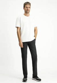 T-shirt bianca a maniche corte, vestibilità comoda, abbinata a pantaloni neri a vestibilità slim. Modello che indossa sneakers grigie, in piedi davanti a uno sfondo semplice.