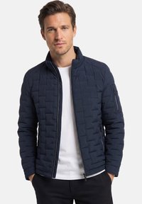 Mann trägt eine marineblaue gesteppte Jacke mit Reißverschluss über einem weißen Hemd, die Hände in den Taschen steckend, steht vor einem schlichten weißen Hintergrund.