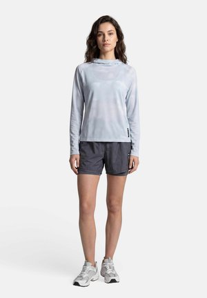 Jeune femme debout, portant un haut à manches longues gris clair, un short gris foncé et des chaussures de sport grises sur un fond blanc uni.