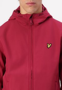 Casaco com capuz vermelho, feito de material suave, com fecho frontal e um logo de pássaro bordado em amarelo no lado esquerdo do peito.