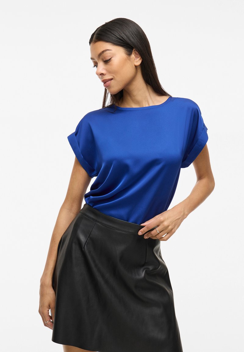 Top blu in satin con maniche arrotolate, scollatura tonda e vestibilità comoda. Abbinato a una minigonna in pelle nera. Tessuto liscio e colore vivace.