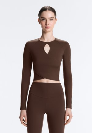 Tânără purtând un top crop maro cu mâneci lungi și decupaj frontal, potrivit pe corp, și colanți asortați cu talie înaltă, stând pe un fundal simplu.
