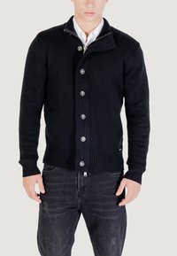 Uomo che indossa un cardigan nero a bottoni sopra una camicia bianca con colletto e jeans neri sbiaditi, in piedi contro uno sfondo chiaro e semplice.