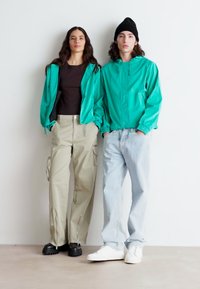 Două persoane poartă jachete verzi strălucitoare și colorate. Femeia are pantaloni cargo bej, în timp ce bărbatul îi asortează cu blugi decolorati și adidași.