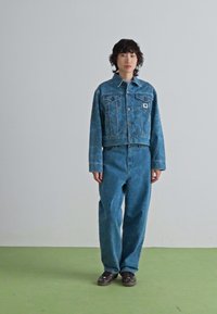 Personne debout sur un sol vert contre un mur clair uni, portant une veste en jean bleue à motifs et un jean ample assorti avec des baskets noires.