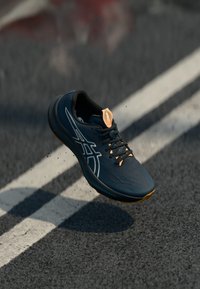 Chaussure de sport de couleur bleu marine foncé avec une surface texturée, des accents blancs et un logo orange au talon. Design léger avec des lacets.