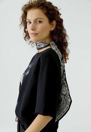 Frau mit lockigem braunem Haar, die ein schwarzes Hemd und einen schwarz-weiß gemusterten Schal trägt, der um ihren Hals gebunden ist, vor einem schlichten Hintergrund.