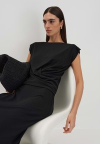 Abito nero con maniche corte, tessuto drappeggiato e una superficie liscia. Accentuato da una borsa a tracolla nera intrecciata. Modella seduta su una sedia bianca.
