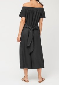 Jumpsuit nero off-shoulder con corpetto smockato, legatura in vita, gambe ampie e texture liscia. Indossato con sandali.