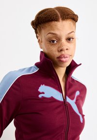 Bordeaux kleurige ritsjack met blauwe accenten en witte strepen, met een groot roze Puma-logo op de borst. Glad stoftextuur, ontwerp met hoge kraag.