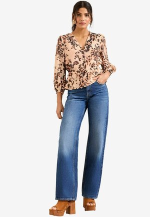 Helle beige Bluse mit schwarzem Leopardenmuster, V-Ausschnitt und Taillierung mit Knoten, kombiniert mit hochgeschlossenen blauen weit geschnittenen Jeans und braunen Plateauschuhen.