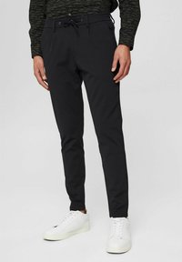 Muž v slim černých kalhotách s ná DRAWSTRING a bílých teniskách, stojící na jednobarevném světlém pozadí.