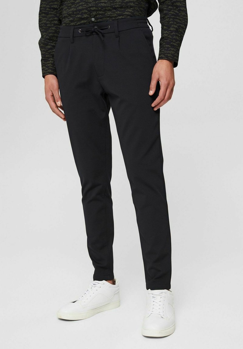 Muž v slim černých kalhotách s ná DRAWSTRING a bílých teniskách, stojící na jednobarevném světlém pozadí.