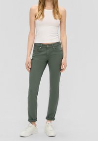 Pantalones ajustados verde oliva con textura suave, cinco bolsillos y dobladillo enrollado, combinados con una camiseta de tirantes blanca acanalada y zapatillas blancas.