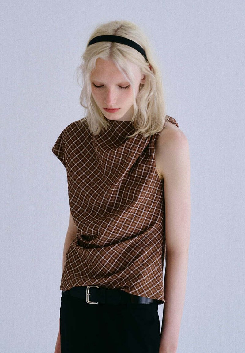 Jeune personne aux cheveux blonds pâles et un bandeau noir porte un haut marron à carreaux sans manches et un pantalon noir avec ceinture, regardant vers le bas.