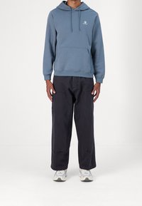 Sudadera con capucha de color azul claro, con bolsillo frontal y logo blanco, combinada con pantalones negros holgados y zapatillas blancas. Ajuste casual y relajado.
