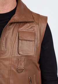 Braune Lederweste mit hohem Kragen, Frontreißverschluss und zwei Taschen. Verfügt über einen kleinen Ring und eine strukturierte Oberfläche. Getragen über einem schwarzen Hemd.