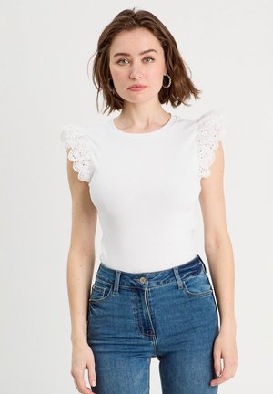 Donna con capelli castani corti che indossa una canotta bianca senza maniche con spalle arricciate in pizzo e jeans blu a vita alta su uno sfondo semplice.