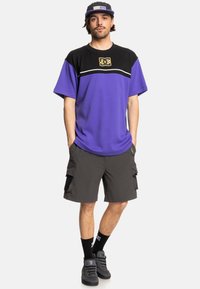 Fialová a černá košile s krátkým rukávem se bílým pruhem, v kombinaci s tmavými cargo šortkami. Nosené s černými ponožkami a šedými botami.