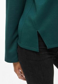 Top de manga larga en color teal con un diseño de dobladillo dividido, que cuenta con una textura suave y detalles de costura, combinado con jeans negros ajustados.