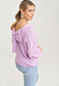 Liliowy sweter z dzianiny o szerokim dekolcie i kokardką z tyłu. Połączony z jasnoniebieskimi dżinsami z wysokim stanem; o fakturowanym wykończeniu.