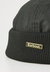 Fekete bordázott kötött sapka sima szövettel a tetején, arany szegésű fekete címkével, amelyen a "Barbour" márkaneve szerepel a szegélyen.