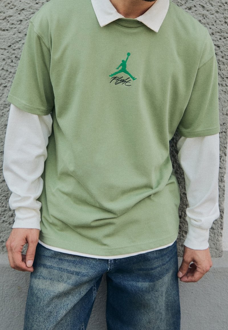 Homme portant une chemise blanche longue superposée sous une chemise à manches courtes vert clair avec un logo "Flight" vert et un jean en denim devant un mur texturé.