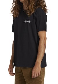 Czarna bawełniana koszulka z krótkim rękawem, luźnym krojem i białym logo na piersi. Noszona z brązowymi spodniami z korduru.