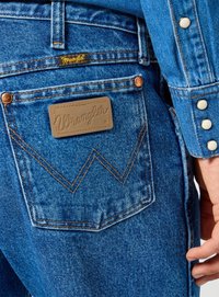 Takapocketi Wrangler sinisissä farkuissa, joissa on nahkainen logo, näkyvää oranssia ommelta ja osa nuppineulaverkko-takkihuputta.