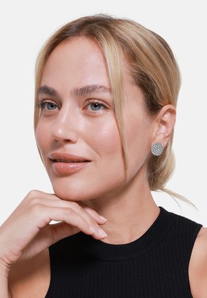 Sølv stiftøredobber med et teksturert, vevd mønster. Båret av en person med lys hud og rett, blond hår. Svart ribbet topp er synlig.