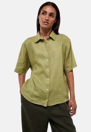 Femme portant une chemise boutonnée à manches courtes vert olive clair et un pantalon foncé, debout les mains dans les poches devant un fond uni.