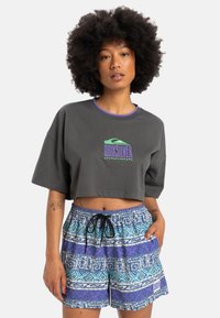 Grå croppete t-skjorte med lilla kant og logo foran, kombinert med blå og lilla mønstrede shorts med elastisk midje og snøring.