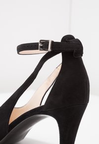 Chaussure à talon haut en daim noir avec bride en T, boucle à la cheville et arrière arrondi, présentée sur un fond clair uni.