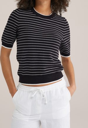 Woman wearing a short-sleeve black knit top with white horizontal stripes and white drawstring pants, standing with hands in pockets.