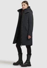 Manteau long noir avec fermeture éclair, capuche ajustable et plusieurs poches. Associé à des bottes noires à lacets montant jusqu'au genou et à des leggings.