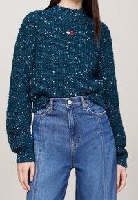 Teal gespikkelde gebreide trui met het "Tommy Jeans" logo, gecombineerd met hoge blauwe denim jeans op een staande persoon.