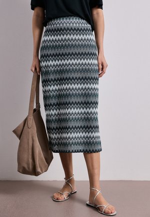 Kobieta w czarnym topie i ołówkowej spódnicy midi w zygzakowaty wzór, trzyma beżową zamszową torbę typu tote, nosi srebrne płaskie sandały na paski.