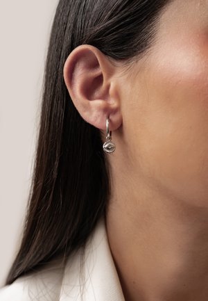 Nahaufnahme eines Frauenohres mit glattem, dunklem Haar, das einen kleinen silbernen Creolenohrring mit einem hängenden runden Anhänger mit einem Augendesign trägt.