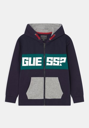 Sweat à capuche zippé bleu marine avec poches avant grises, bande poitrine verte arborant le texte blanc "GUESS?", cordons de serrage rouges à l'intérieur de la capuche.