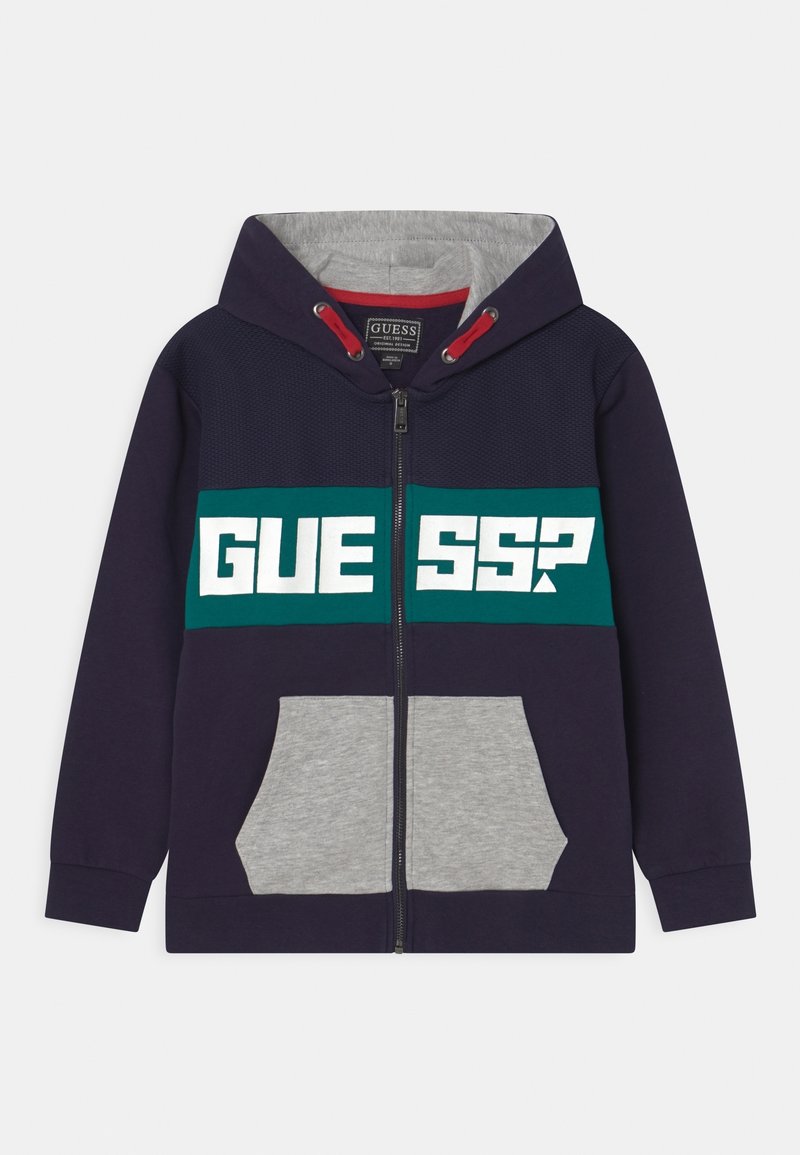 Sweat à capuche zippé bleu marine avec poches avant grises, bande poitrine verte arborant le texte blanc "GUESS?", cordons de serrage rouges à l'intérieur de la capuche.